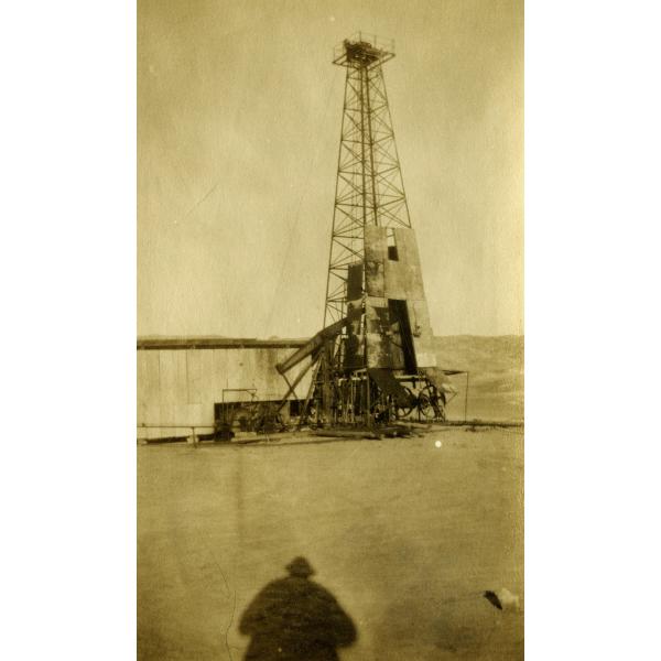 An oil derrick covered halfway up in metal siding. There is mechanical equipment on the right. The ground is flat dirt and there is a hill in the background. The photographer's shadow is at the front of the photo.