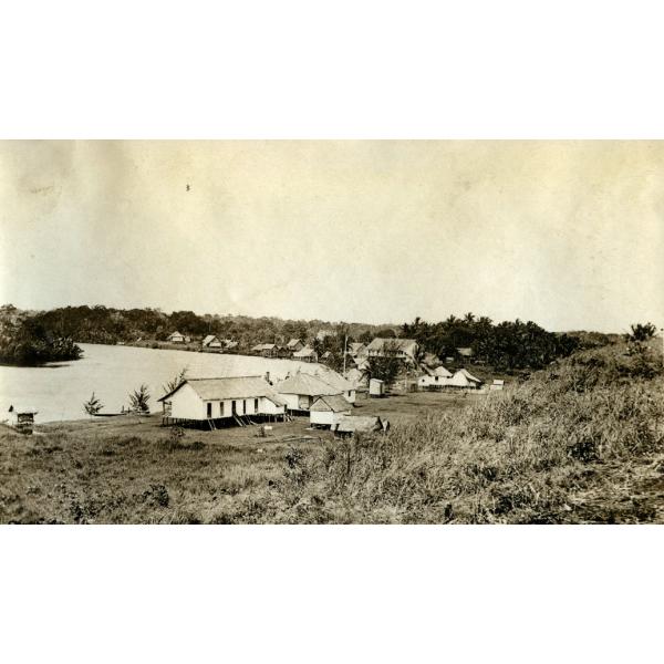 A photo of a group of buildings on stilts along the edge of a river. There is a cluster of trees on the other side of the river and a hill with grass in the foreground.