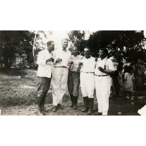 A photo of five men wearing white shirts stand together, holding small glasses of alcohol. There is a group of people behind them to the right.