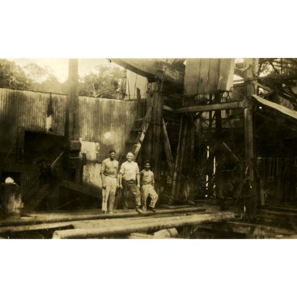 Three men standing in front of an oil rig covered with metal siding. They are standing on a pile of casing.