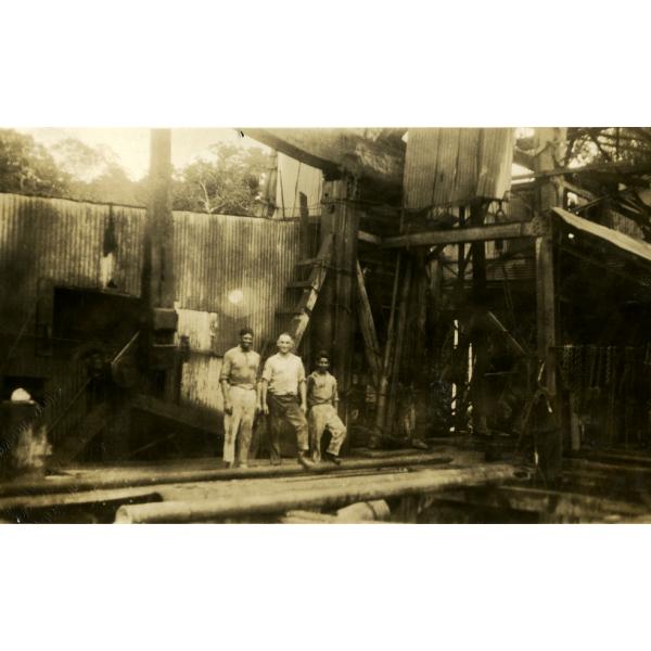 Trois hommes debout devant une installation de forage pétrolier recouverte d’un revêtement métallique. Ils sont debout devant une pile de tubage. 