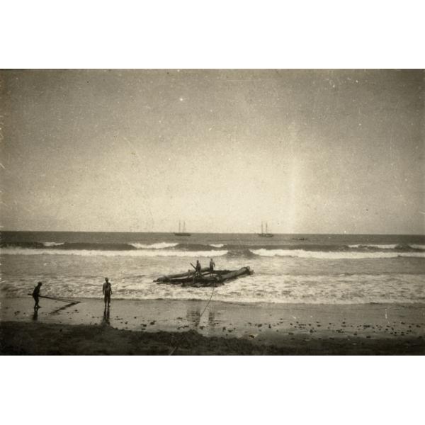 A photo of two men standing on a raft in shallow water and two on the beach. There are two ships anchored off-shore. 