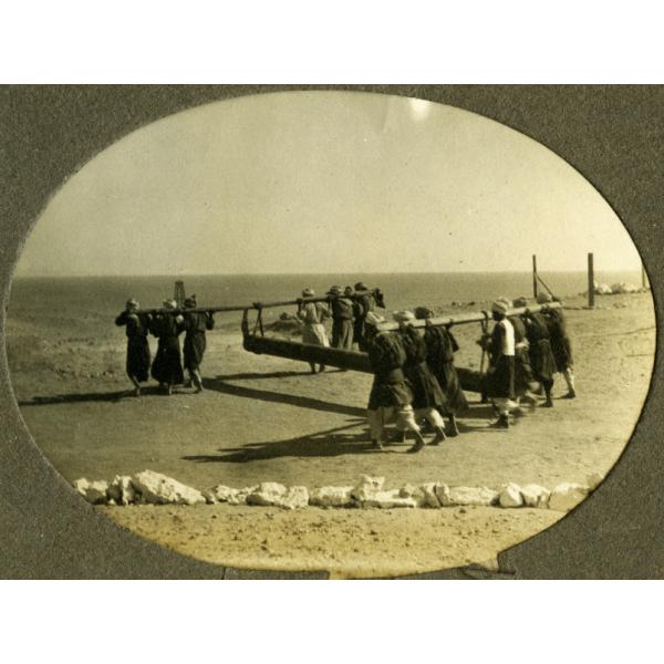 Une photo d’un groupe d’hommes transportant un tuyau. La conduite est fixée à deux poutres et les hommes se tiennent debout à l'avant et à l'arrière de la conduite et soutiennent la poutre sur leur dos. Le sol est plat et il y a une rangée de rochers blancs à l’arrière-plan. 