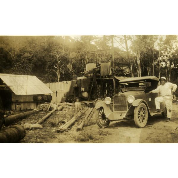 Fred Webb standing with one foot on the step of a car door. He is wearing white with a pith helmet. There is an oil rig with four legs behind him and a building with a roof on the left.