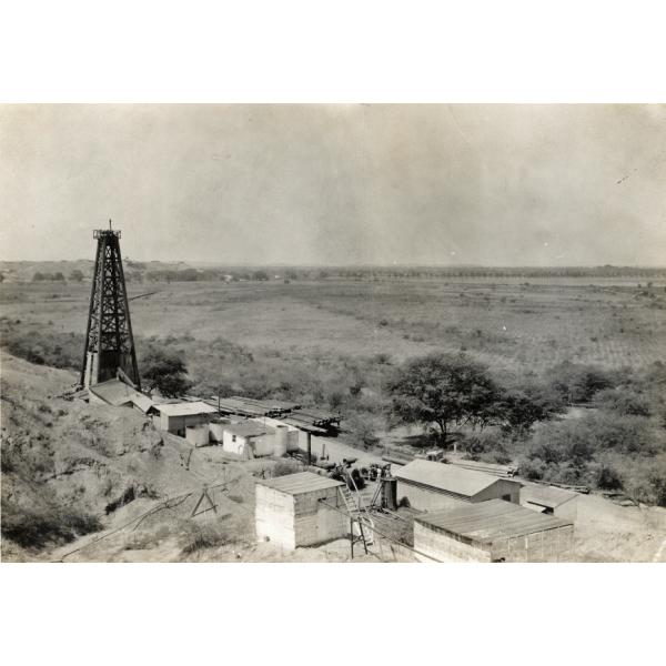 A single oil rig in an open plain with piles of timber and casing beside a few small buildings. 