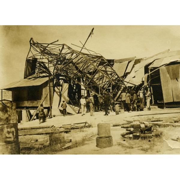 A photo of a metal derrick that has fallen over on top of a structure with a metal roof and siding. A group of people are standing in front of it looking at the damage. There are metal cylinders and casing in the foreground.