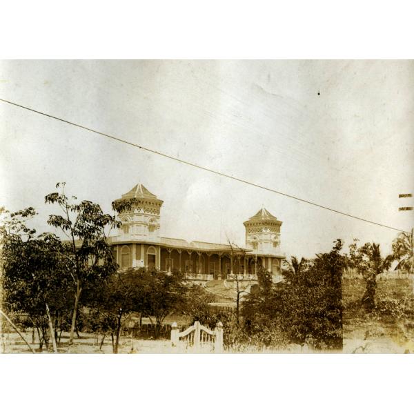 A photo of a building in behind trees and a gate. There are turrets on the corners of the building that are visible.