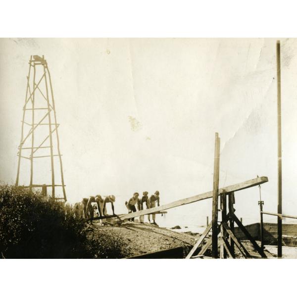 Une photo de six hommes appuyant sur une traverse en bois soutenue par un cadre en bois. Il y a une structure en bois à quatre mâts derrière eux et un buisson sur la droite. 