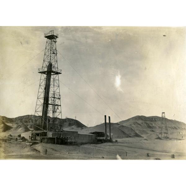 An oil rig with four legs and crossbeams for support. There are four lines attaching it to the ground and there is a covered building at its base. There are dirt hills and another rig in the background.