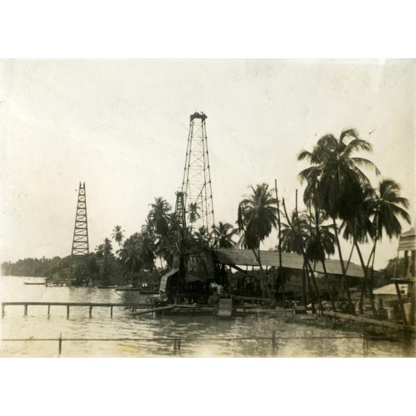 Two oil rigs beside the water. There are three docks coming out from the shore line and palm trees along the edge of the water. There is a wooden roof over a large wheel beside the rig on the right.