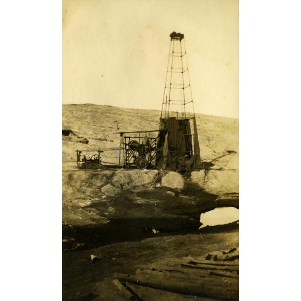 A metal oil rig. The base is covered in dark cloth. There is a roof over the wheels and belts beside the rig. There is casing on the ground in front of the rig and a bare hill in the background.