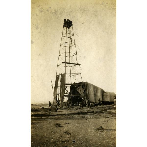 An oil derrick with four poles. The pole on the right is bent out of shape. There are three men on the top of the derrick and another climbing towards the, There is a group of men at the bottom of the rig, and part of the rig is covered in dark cloth.