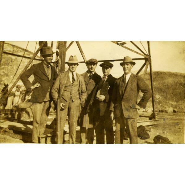 A photo of five International Drillers standing at the base of an oil derrick. They are wearing suits and hats.