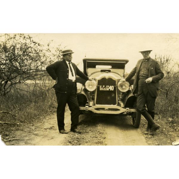 Webb et Burns debout de chaque côté d'une voiture stationnée sur un chemin de terre. Il y a de petits arbres de chaque côté d'eux. La plaque d'immatriculation entre les phares indique : 1924. I.P.Co. STA.ELENA. 40 ».  