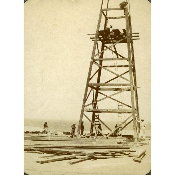 A wooden oil derrick with four legs. A group of men stand at its base surrounded by a pile of wooden planks. Three men are on the derrick and there are two ladders resting against the lowest support beam.