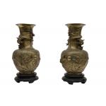 A pair of brass urns sitting on wooden bases. Each vase has a design of two dragons or serpents that is raised off of the urns. There is a serpent wrapped around the neck of both.