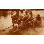 A photo of a group of men crouching with an alligator on a river bank. Its mouth is open, showing its teeth. 