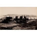 Four large, dark refinery stills with pipes standing along them. There are round storage tanks behind them and hills in the background. 