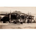 A photo of a single-story buildings with latticework on the porch and palm trees in front. There are two cars parked outside beside a dirt road. Hydro wires run overhead. 