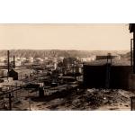 A photo of an oil refinery. There is a dark storage tank with stairs leading to the top on the right. There are buildings with pipes running between them in the middle and a group of light-coloured storage tanks at the back. There are hills in the background. 