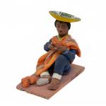 Figurine d’un homme mangeant un fruit et portant des sandales, un tailleur-pantalon bleu, une écharpe jaune, ainsi qu’un poncho orange drapé sur une épaule. Il porte un chapeau jaune inversé et quatre objets orange en forme d'œuf sont à ses pieds.