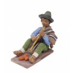 Figurine d’un homme jouant de la flute et portant des sandales, des pantalons verts, une chemise bleue, ainsi qu’un poncho brun sur une épaule. Il porte un chapeau gris et trois objets orange en forme d'œuf sont à ses pieds. 