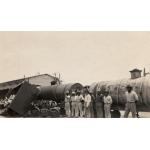 A photo of a group of men looking at a railway carriage that was derailed. There are two cylindrical carts still on the tracks and a building in behind. 