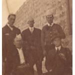 Five International Drillers, three standing and two sitting. They are wearing dark suits. Four of the men are wearing paper hats and one (bottom right) is wearing an ascot cap.  