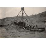 A group of men are putting together a bulldozer using three poles tied together at the top with a chain coming down from the middle to help lift the heavy pieces. There is a hill dotted with trees in the background. 