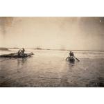 Trois hommes debout sur un radeau en rondins et trois hommes à bord d’un canot. Il sont en eau peu profonde et des petites vagues se dirigent vers eux. Il y a deux bateaux sur l’eau et en arrière-plan.