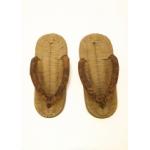 A pair of light brown flat thong sandals. Rice straw has been woven together to create the bed of the shoes and a piece of brown material forms the thong. It is attached to the front in the centre and two-thirds of the way back to both sides. 