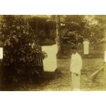 A photo of a man standing in front of white pillars topped with black on a fence. There is a building with a porch in the background. The man is wearing a white outfit with a dark hat.  