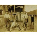 Three men in light shirts and dark pants, holding guns, in front of a wooden picket fence. The man in the centre is sitting on the ground and the other two are standing. 