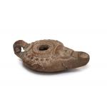 Side view of the terracotta oil lamp. There is a handle at one end and a spout at the other that has a face on it. There is an opening in the middle of the lamp surrounded by detailing. 