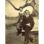 A woman in a long, dark dress and a man in a dark suit and tie sit on wooden planks beneath a tree branch.