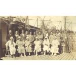 A photo of two rows of people on a wooden deck. The men are standing in the back row, wearing suits. The women are sitting in chairs in the front, wearing dresses and hats. 