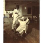 A woman lying back in a wicker reclining chair, wearing a white dress and hat. A man in a white suit stands behind her. They are on a ship deck with a wooden floor.