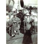 A man and a woman sitting on the deck of a ship. There are life boats hanging to the left and a palm frond behind them. The floor is made from wooden boards.