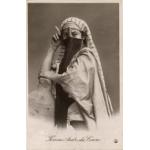 Une carte postale avec une femme voilée portant une étoffe rayée blanche sur la tête. Elle porte deux bracelets, un sur chaque poignet, et tient une cigarette. L’inscription au bas indique : « Femme arabe du Caire »  