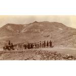 A photo of Egyptian workers standing in a line, hauling a boiler with ropes along a road in the desert. The boiler has four weeks and is has two cylindrical components put together. 