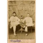 A photo of two men wearing white, sitting cross-legged on chairs on either side of a small table covered with a cloth. There is a vase of flowers on the table. Both men have a cane in their right hand and are balancing pith helmets on their left knee. There is a painted background of temples in the background.