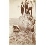 A photo of three men stand on top of a large rock. The man on the right is holding a fishing spear, the man in the middle a gun, and on the left a survey rod. The tide has gone out and mud can be seen under a shallow layer of water.