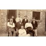 A photo of eight men in front of a stone building with a wooden-framed door and window. Four are standing at the back, three are sitting in chairs, and one is sitting on the ground.