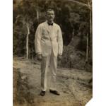 A photo of William O. Gillespie in a white suit with a dark bow tie and shoes. He is standing on a path in front of trees. 