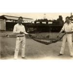 Irwin and Lovelady holding a boa constrictor in between them. They are standing on a dirt road and there are buildings behind them. They are wearing white shifts and pants. 