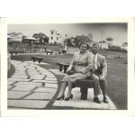 Une photo de Vola et John Braybrook assis sur un banc de parc. Il y a un sentier avec de grosses pierres à leurs côtés et une série de bancs de parcs le long du sentier derrière eux. Elle porte une jupe, un blouson et des talons hauts, et il porte un complet avec une cravate.