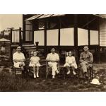 Trois hommes et deux filles (en alternance) sont assis sur des chaises à l'extérieur d'un bâtiment sur pilotis. Ils portent des vêtements pâles. Le bâtiment possède un porche avec des escaliers, ainsi que des balustrades et des poteaux blancs et foncés. 