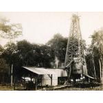 A photo of five men standing by a pile of casing in front of a drilling rig. There is a structure with a roof in the foreground, and a line of trees in the background.