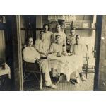 A photo of seven International Drillers around a table indoors with a white tablecloth. They are wearing white and four are sitting on wicker chairs. There is a cup, a pipe, and a bottle on the table.