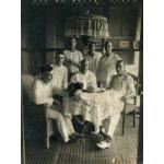 A photo of seven International Drillers around a table indoors with a white tablecloth. They are wearing white and four are sitting on wicker chairs. There is a jar, and a pipe, as well as a dog sitting on the table.